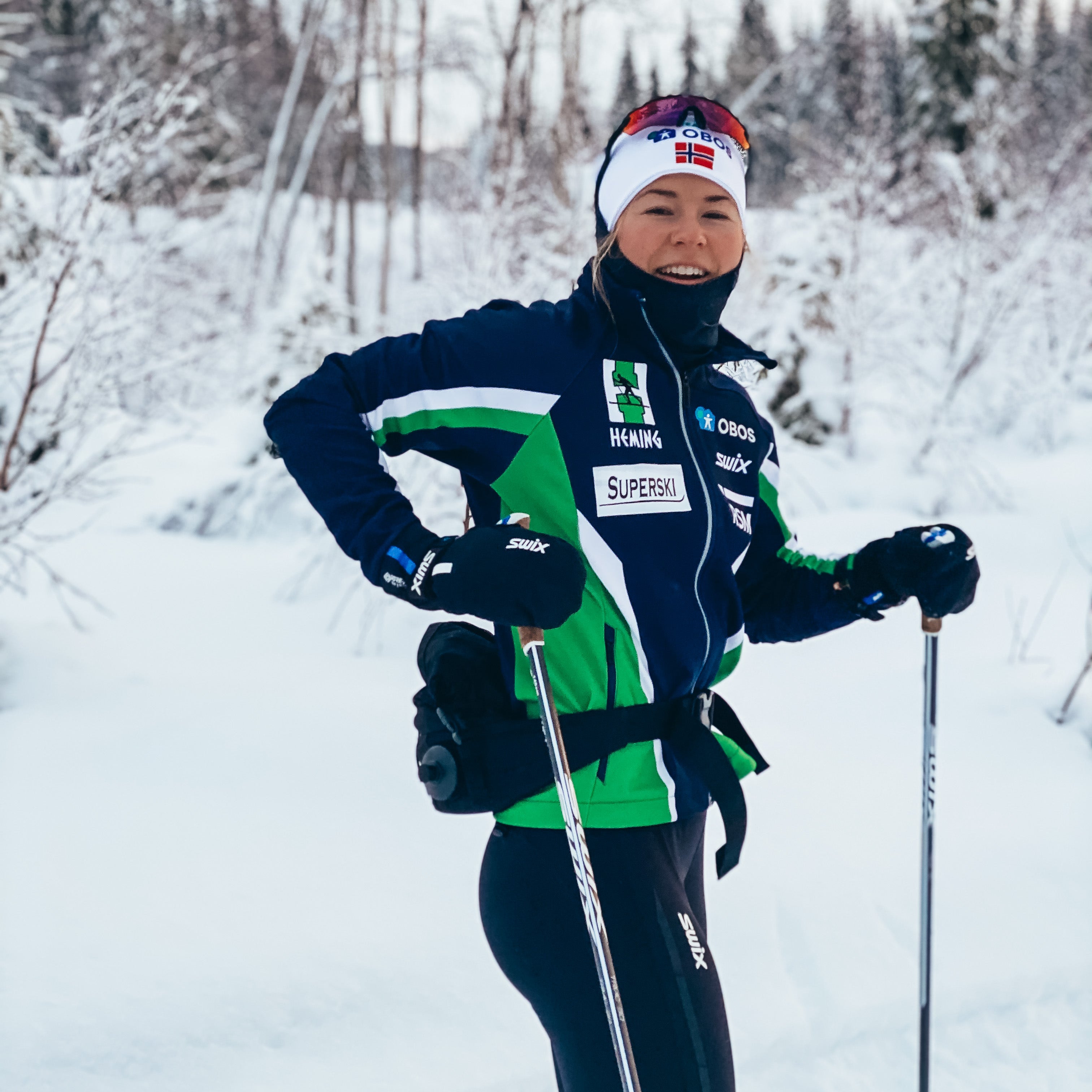 Smilende skiløper i blå og grønn treningsjakke står på langrennsski i snødekt skog.