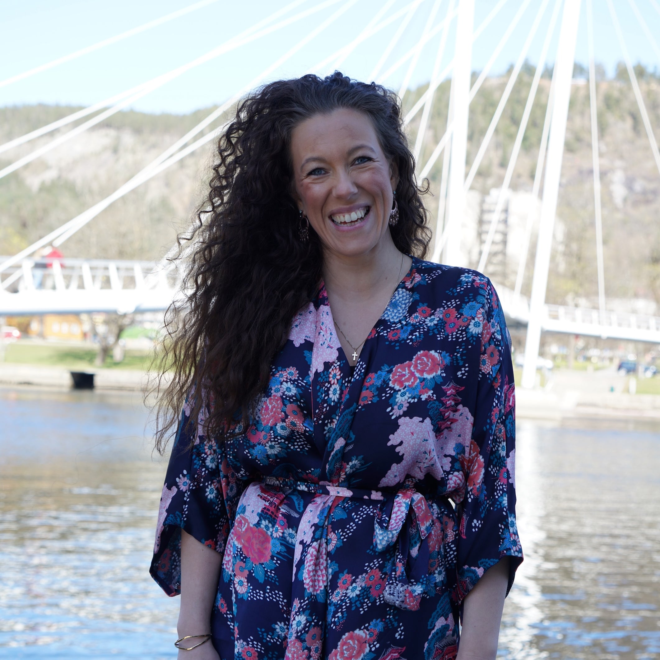 Smilende kvinne med krøllete hår og blomstermønstret jakke foran elv og bro.