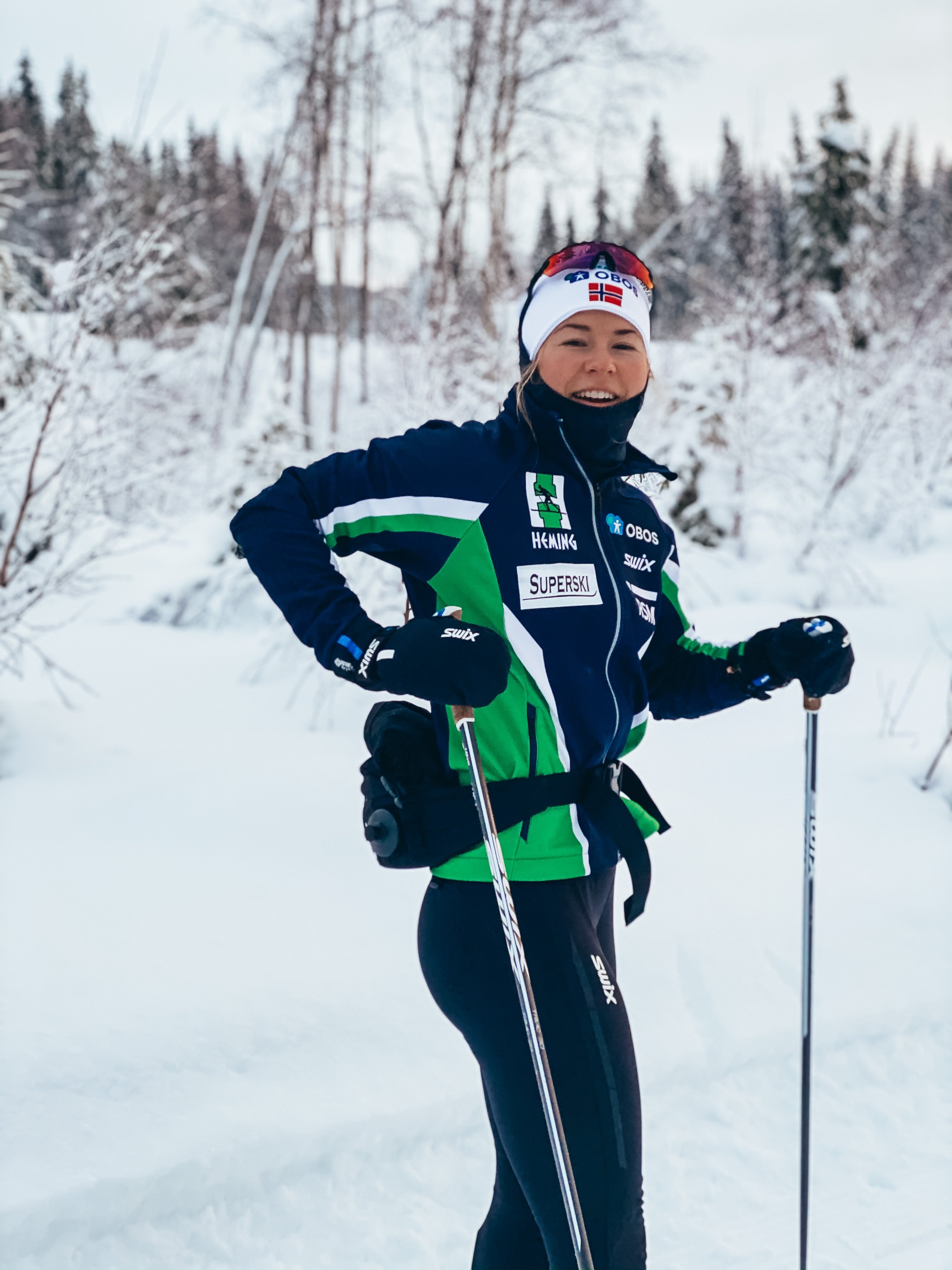 Smilende skiløper i blå og grønn treningsjakke står på langrennsski i snødekt skog.