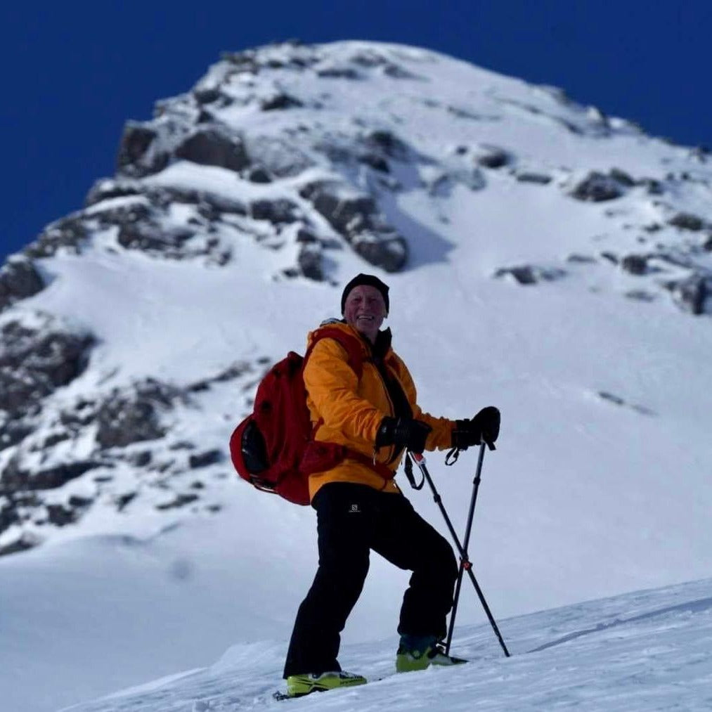 Smilende person i gul jakke og rød sekk står med skiutstyr i snødekt fjellside.