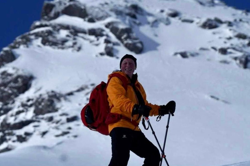 Smilende person i gul jakke og rød sekk står med skiutstyr i snødekt fjellside.