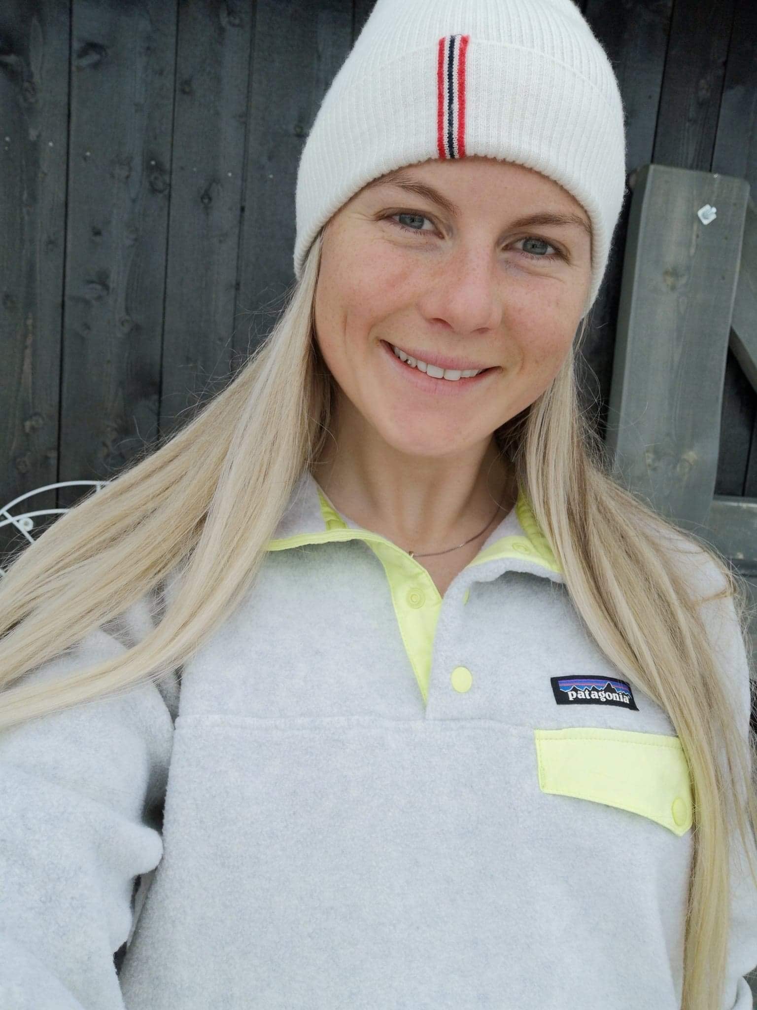 Smilende kvinne med langt blondt hår, hvit lue og grå Patagonia-genser foran mørk vegg.