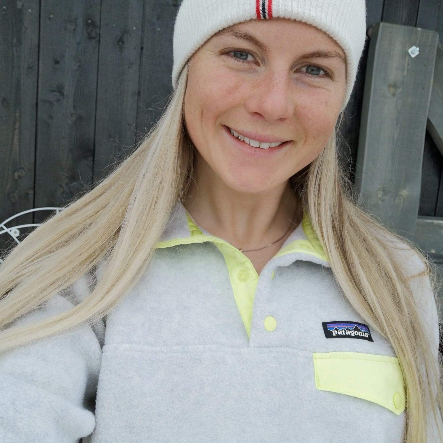 Smilende kvinne med langt blondt hår, hvit lue og grå Patagonia-genser foran mørk vegg.