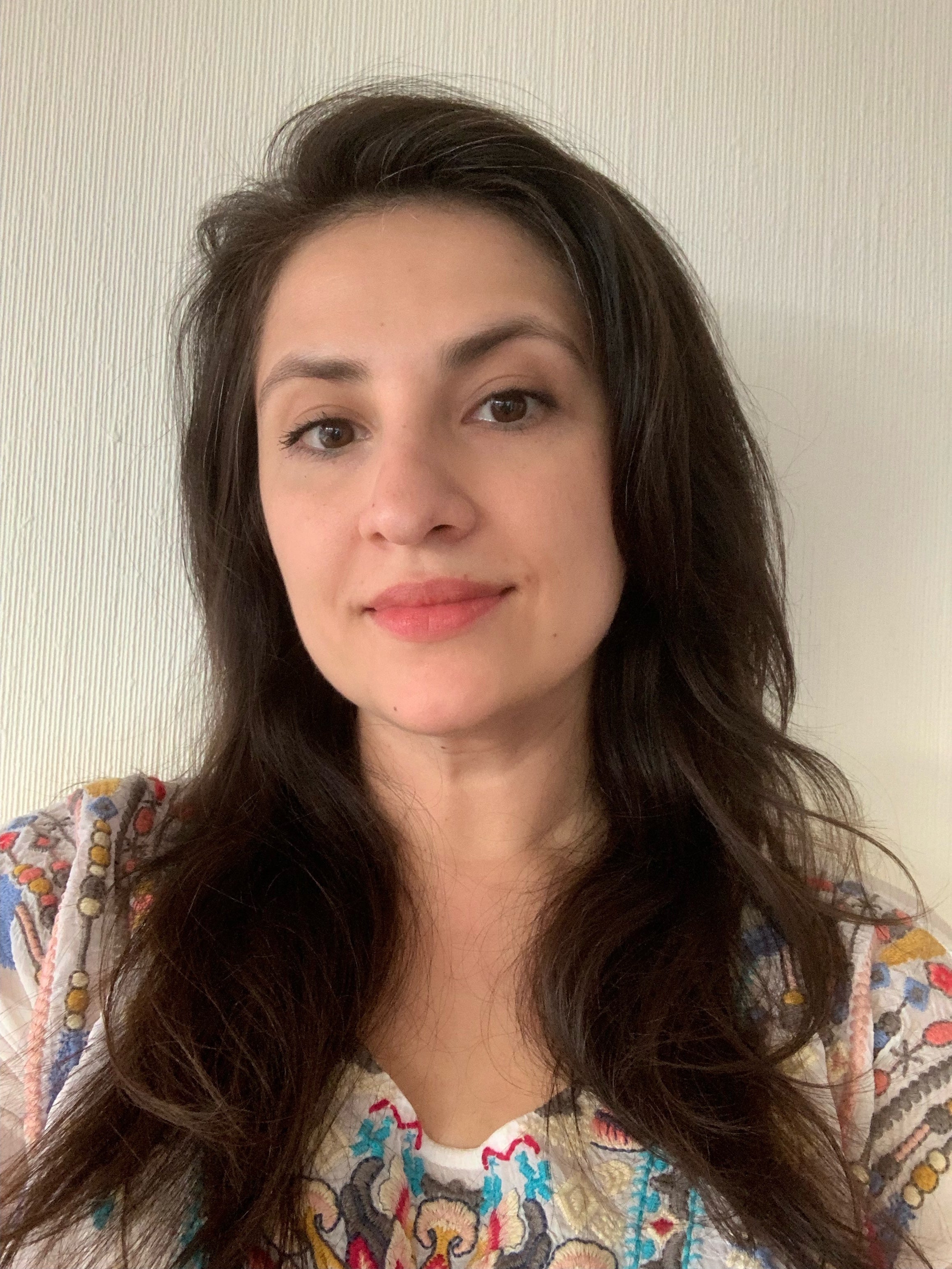 Kvinne med langt mørkt hår og fargerik bluse smiler svakt foran lys vegg.