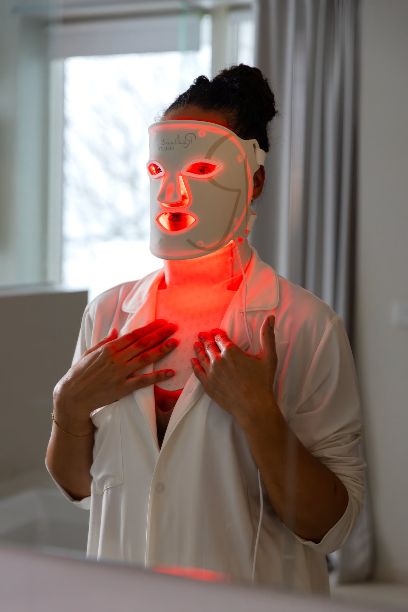 Kvinne bruker LED-ansikts- og halsmaske med rødt lys for hudpleie og lysterapi, i et lyst baderomsmiljø med speil og naturlig lys