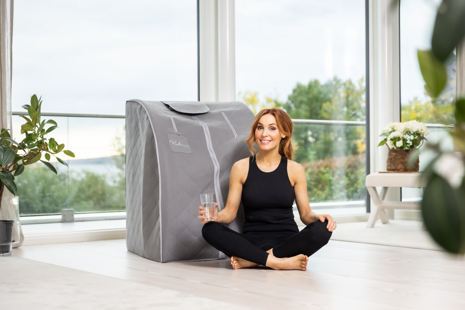 Kvinne i svart treningstøy sitter smilende på gulvet med et glass vann ved siden av en grå IR SaunaPod fra Radiant Health, i et lyst rom med store vinduer og grønne planter.